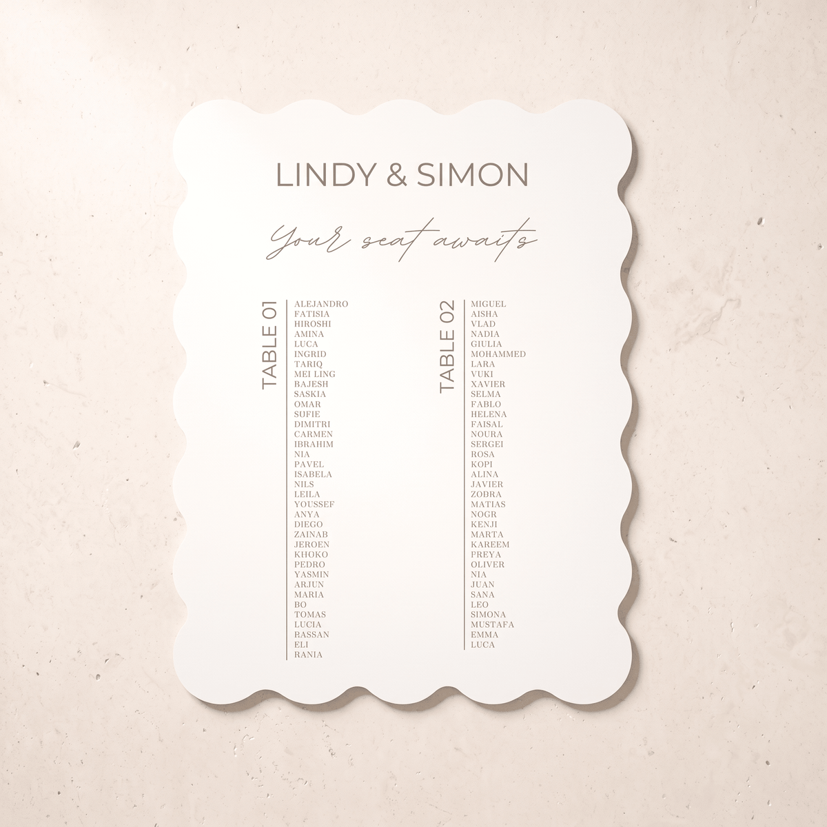 A scalloped-edge wedding seating card with the names 'LINDY & SIMON' and 'Your seat awaits' printed on it, placed on a light beige textured surface.