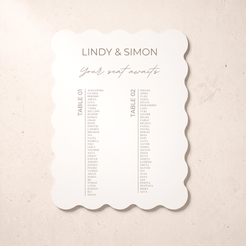 A scalloped-edge wedding seating card with the names 'LINDY & SIMON' and 'Your seat awaits' printed on it, placed on a light beige textured surface.