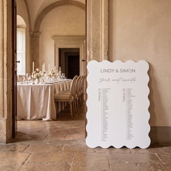 A white scalloped-edge wedding seating chart sign leaning against a stone wall in a warmly lit event venue, with a table setup visible through an open doorway in the background.