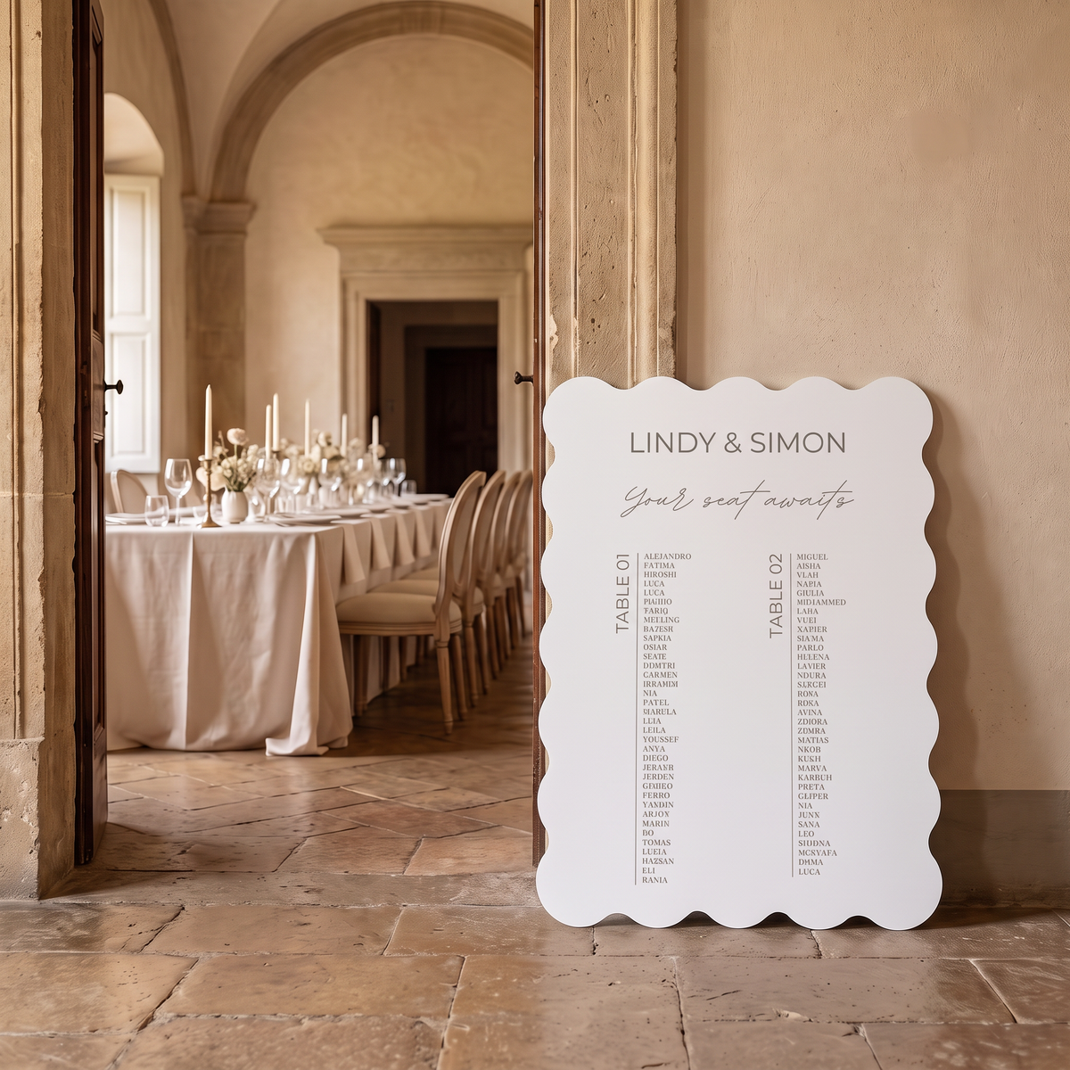 A white scalloped-edge wedding seating chart sign leaning against a stone wall in a warmly lit event venue, with a table setup visible through an open doorway in the background.
