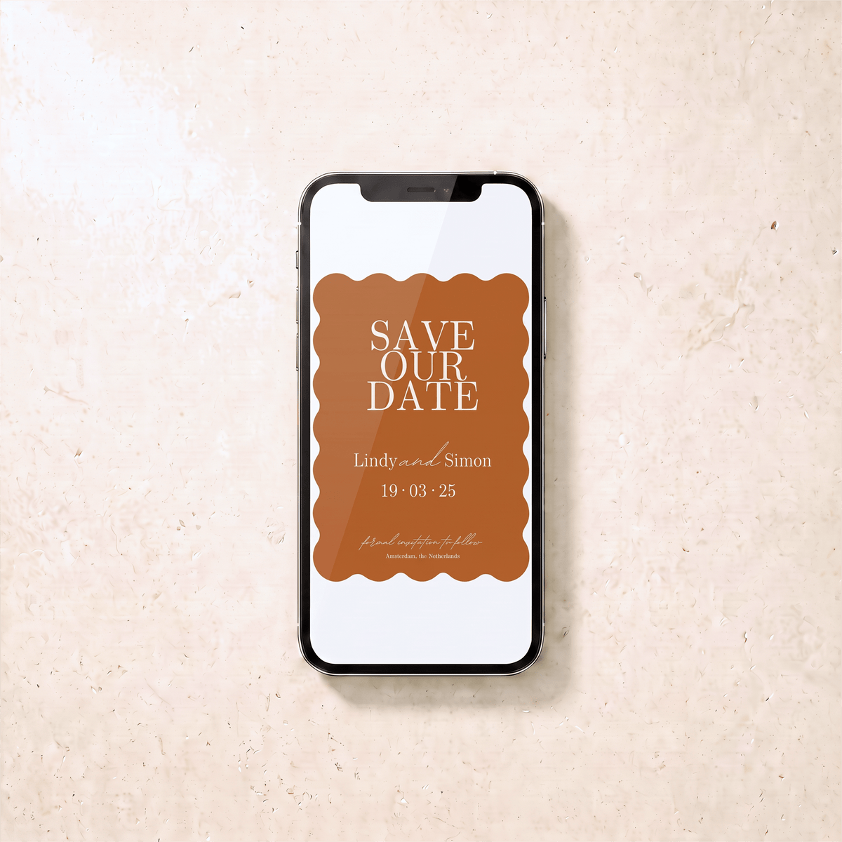 A smartphone lying on a light beige textured surface, displaying a brown and white 'Save Our Date' invitation design with scalloped edges and wedding text.