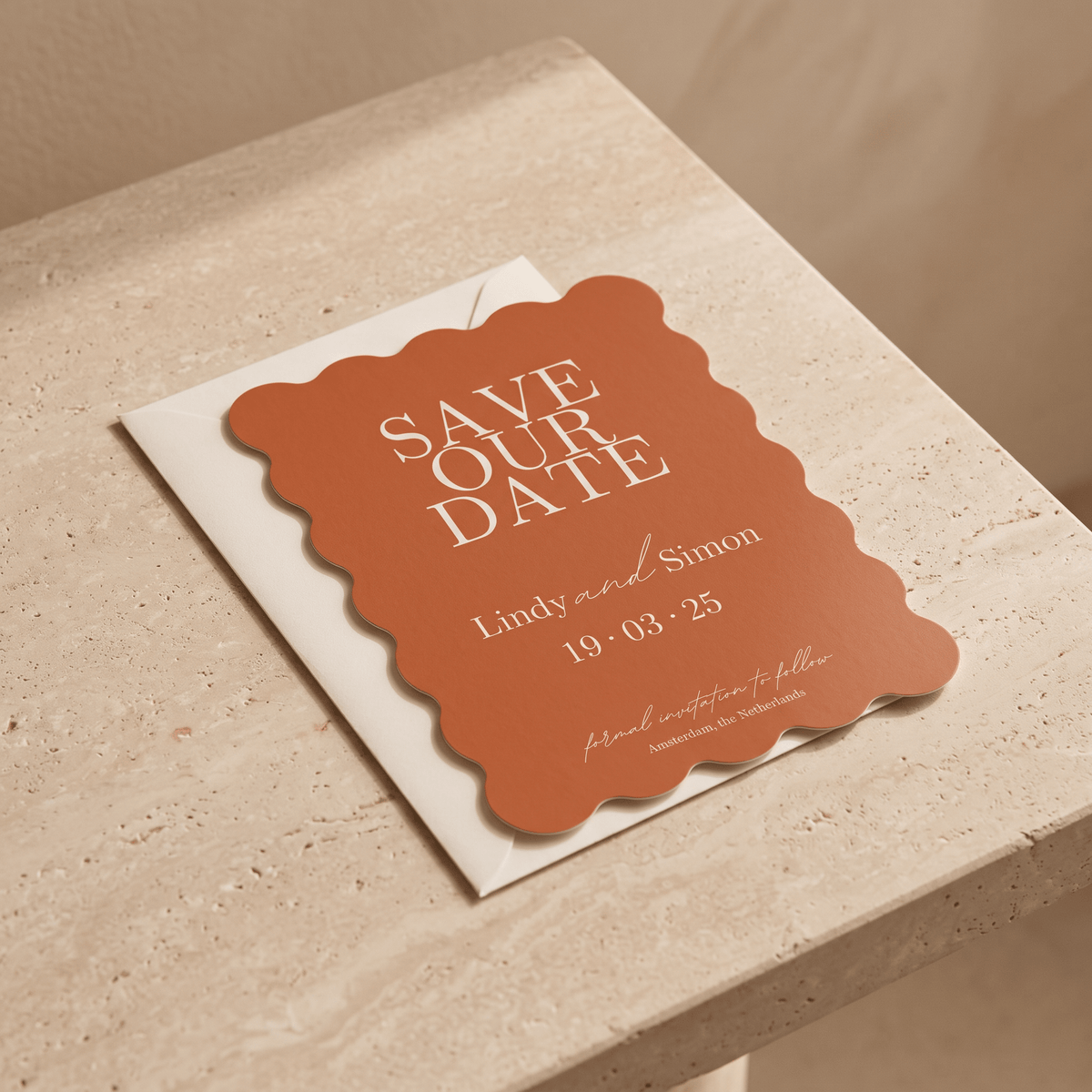 A terracotta-colored scalloped 'Save Our Date' card with white text, resting on a white envelope atop a beige stone surface.