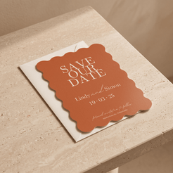 A terracotta-colored scalloped 'Save Our Date' card with white text, resting on a white envelope atop a beige stone surface.
