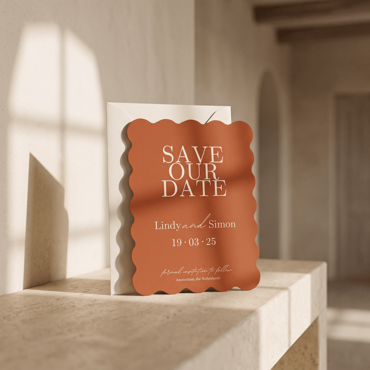A scalloped orange 'Save Our Date' card with white text stands in front of a white envelope on a light stone ledge in a softly lit interior setting.