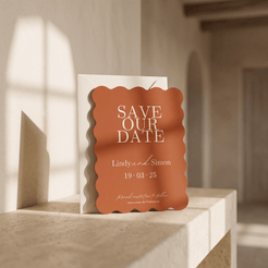 A scalloped orange 'Save Our Date' card with white text stands in front of a white envelope on a light stone ledge in a softly lit interior setting.