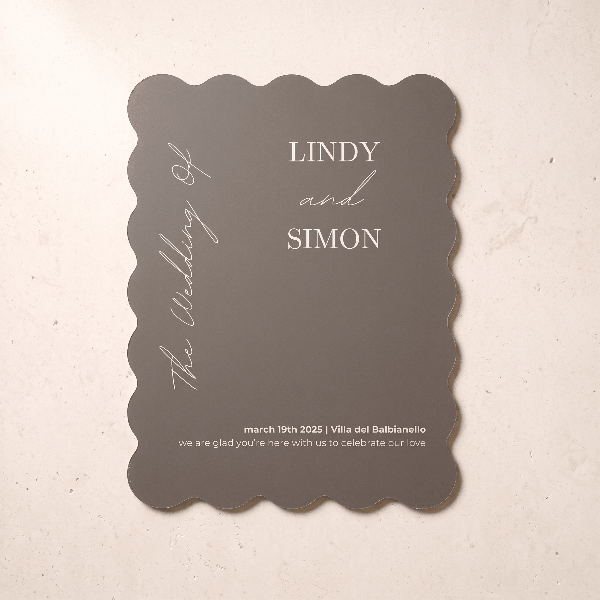 A scalloped-edge wedding invitation card in a muted taupe-gray color with white printed text, displayed flat on a light beige textured surface.