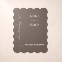 A scalloped-edge wedding invitation card in a muted taupe-gray color with white printed text, displayed flat on a light beige textured surface.