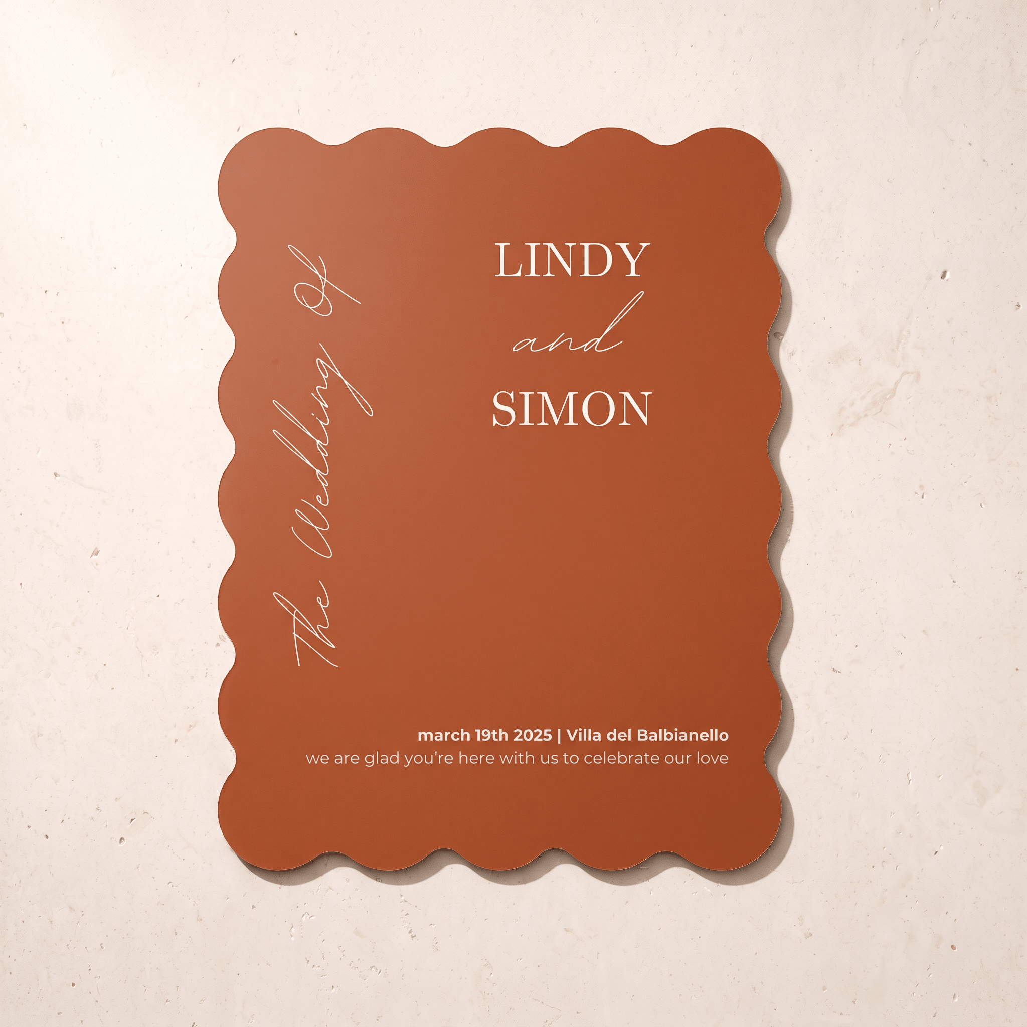 A terracotta-colored wedding invitation card with scalloped edges, featuring white text that reads 'The Wedding of LINDY and SIMON' and event details, displayed on a textured beige background.