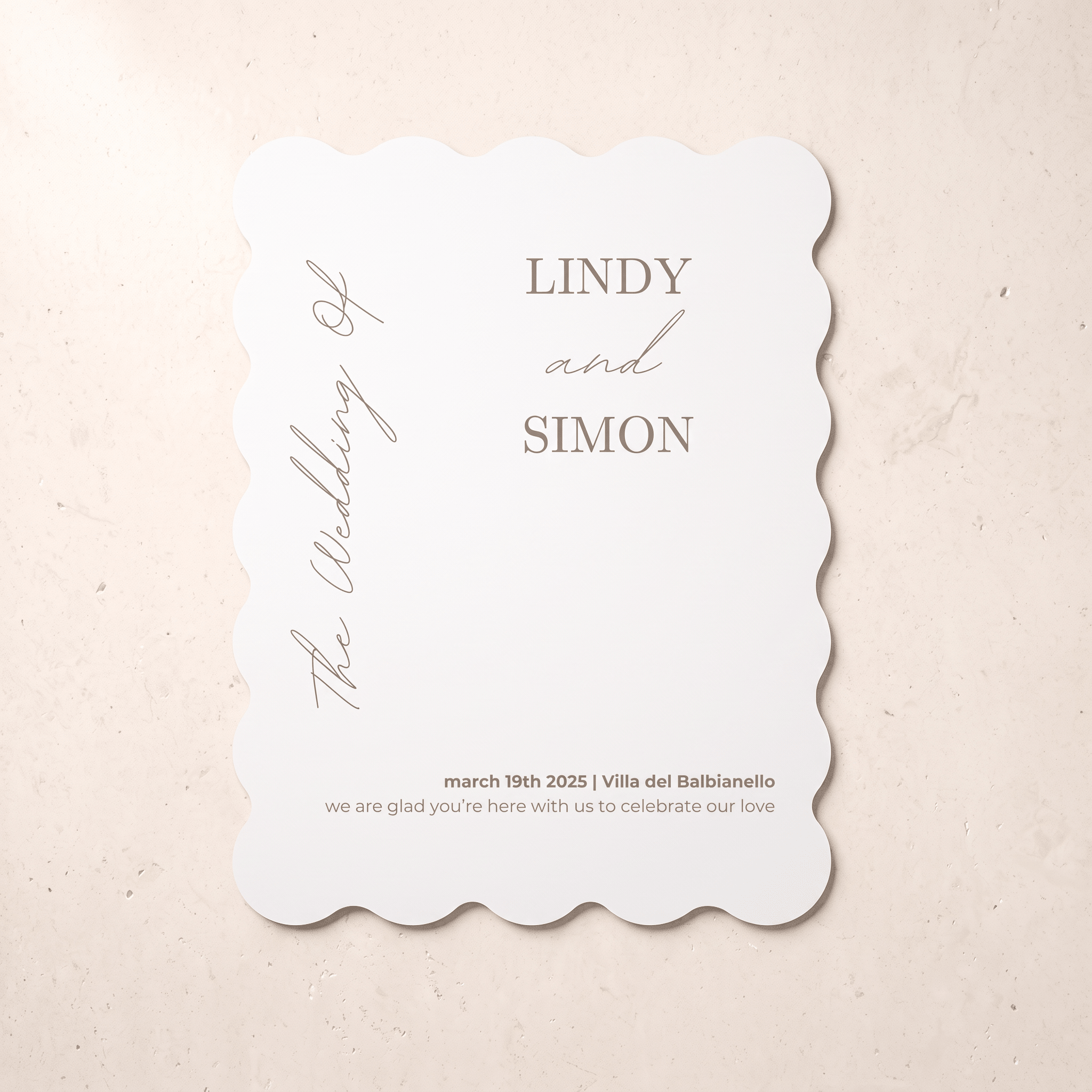A scalloped-edge white wedding invitation card with elegant gray typography, displayed flat on a textured beige surface. The card features placeholder names 'LINDY and SIMON' and wedding details.