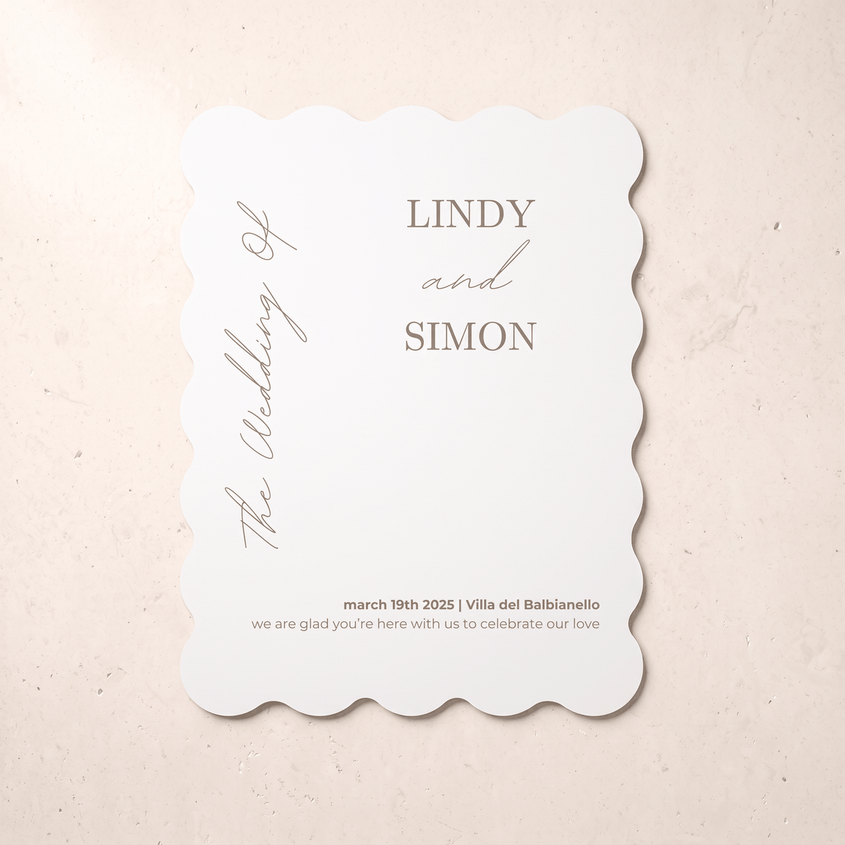 A scalloped-edge white wedding invitation card with elegant gray typography, displayed flat on a textured beige surface. The card features placeholder names 'LINDY and SIMON' and wedding details.