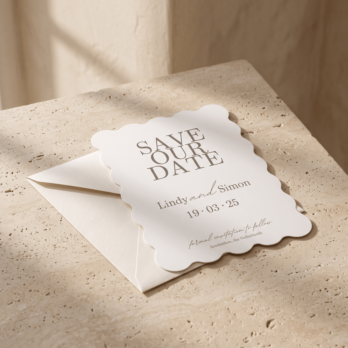 A scalloped-edge white save-the-date card with elegant gray typography, resting on a cream envelope over a textured beige stone surface with soft natural shadows.