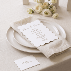A styled table setting with layered white plates, a cream linen napkin, a scalloped white menu card reading 'Time To Dine,' a matching place card reading 'CLAIRE & CO.,' and soft cream flowers in the background on a light beige tablecloth.