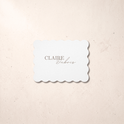 A rectangular scalloped-edge card or label in white with the text 'CLAIRE Dubois' printed in white, placed on a light beige textured surface.