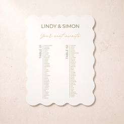 A scalloped-edge wedding seating card with the names 'LINDY & SIMON' and 'Your seat awaits' in olive green and gold typography, displayed on a textured beige surface.