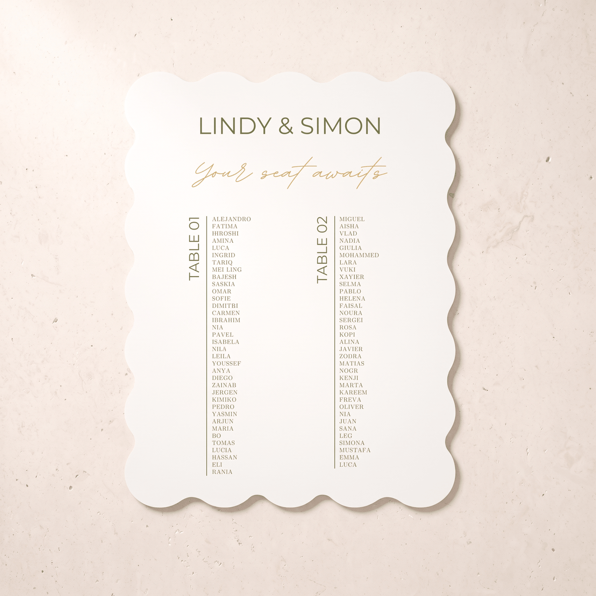 A scalloped-edge wedding seating card with the names 'LINDY & SIMON' and 'Your seat awaits' in olive green and gold typography, displayed on a textured beige surface.