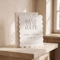 A scalloped white paper save-the-date card with gray text, standing in front of a matching envelope on a stone ledge in a softly lit interior setting.