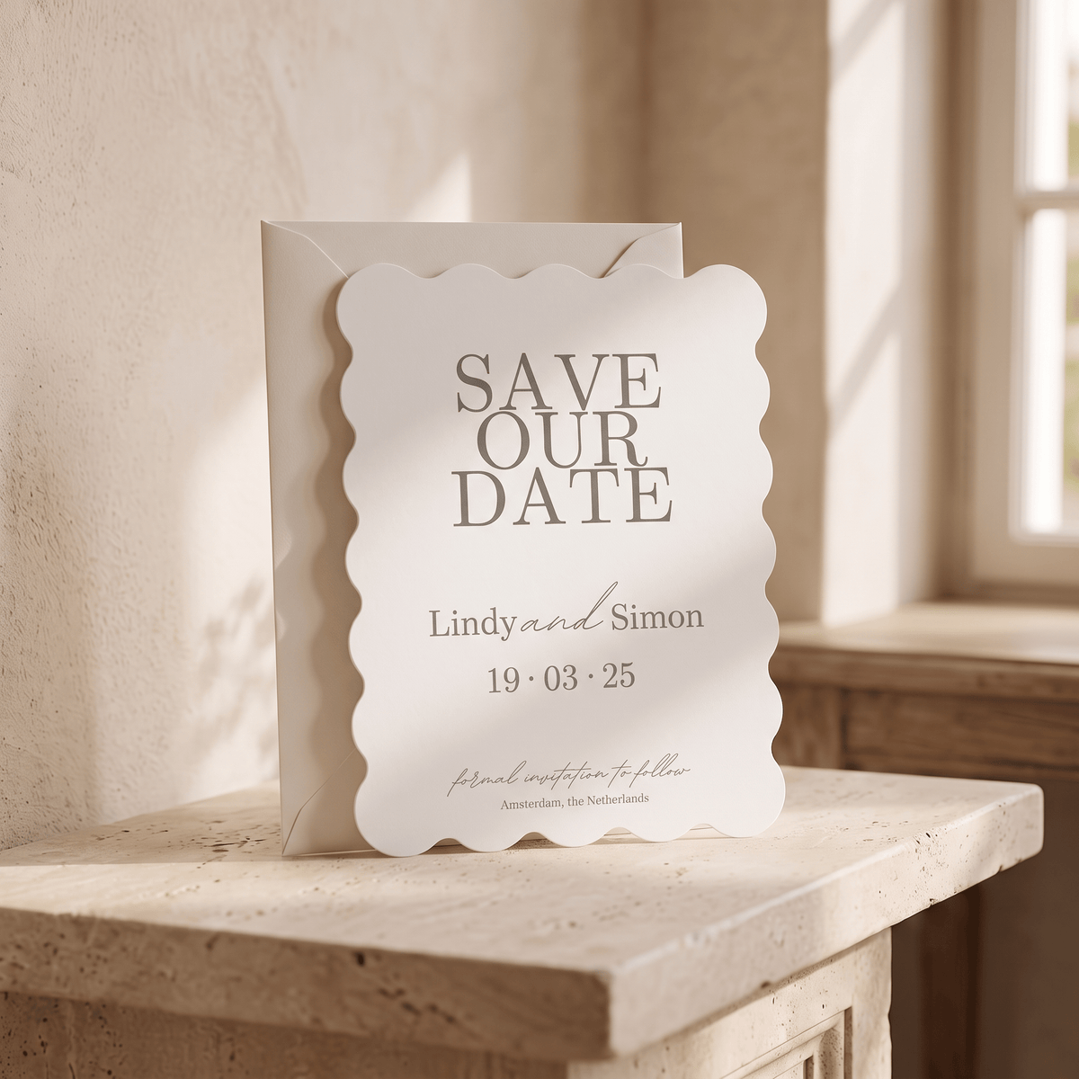 A scalloped white paper save-the-date card with gray text, standing in front of a matching envelope on a stone ledge in a softly lit interior setting.