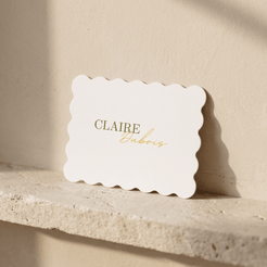 A white scalloped-edge card with the name 'CLAIRE Dubois' printed in gold lettering, leaning against a beige wall on a stone ledge.