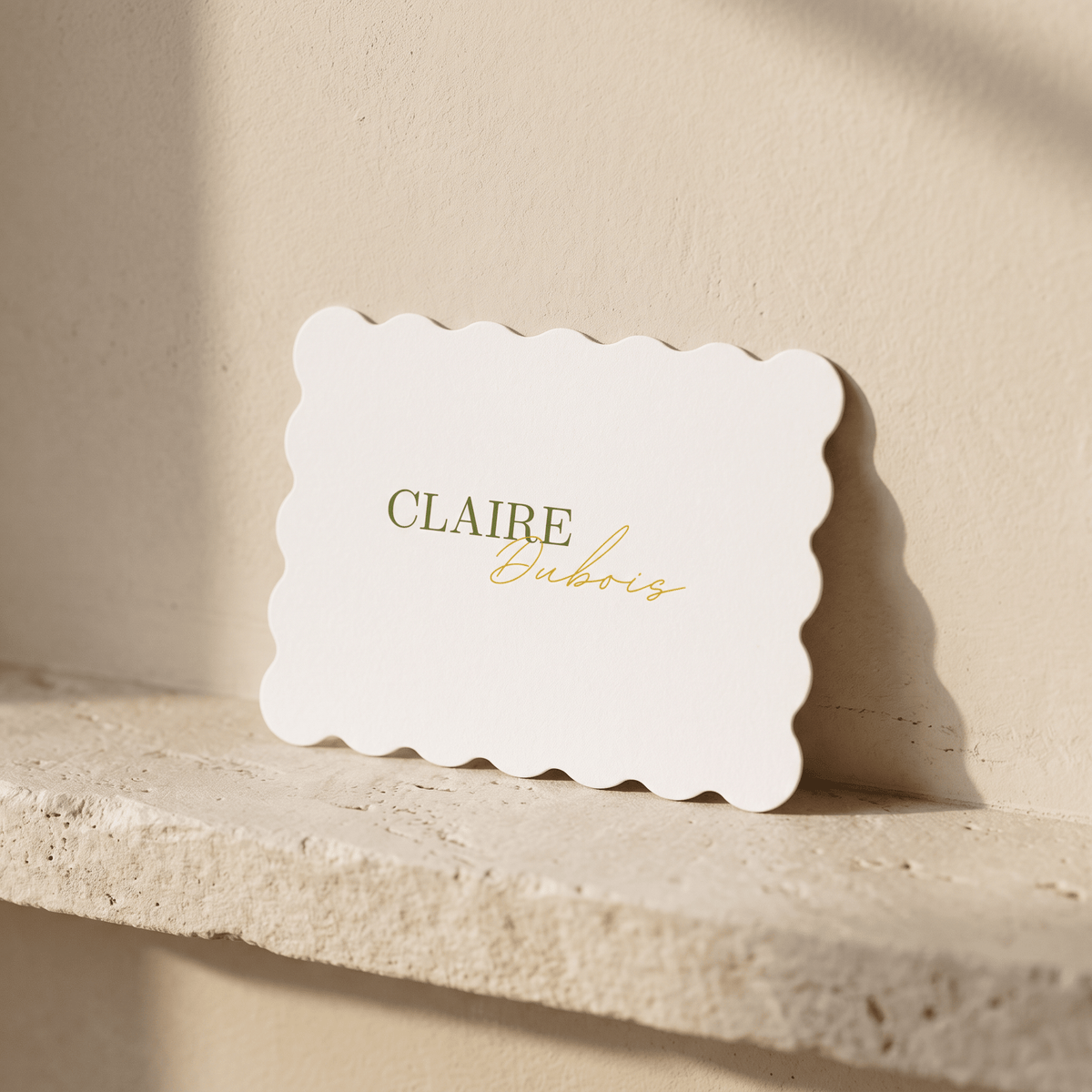 A white scalloped-edge card with the name 'CLAIRE Dubois' printed in gold lettering, leaning against a beige wall on a stone ledge.