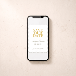 A smartphone lying on a light beige textured surface, displaying a white 'Save the Date' card design with gold typography and scalloped white paper-like borders.