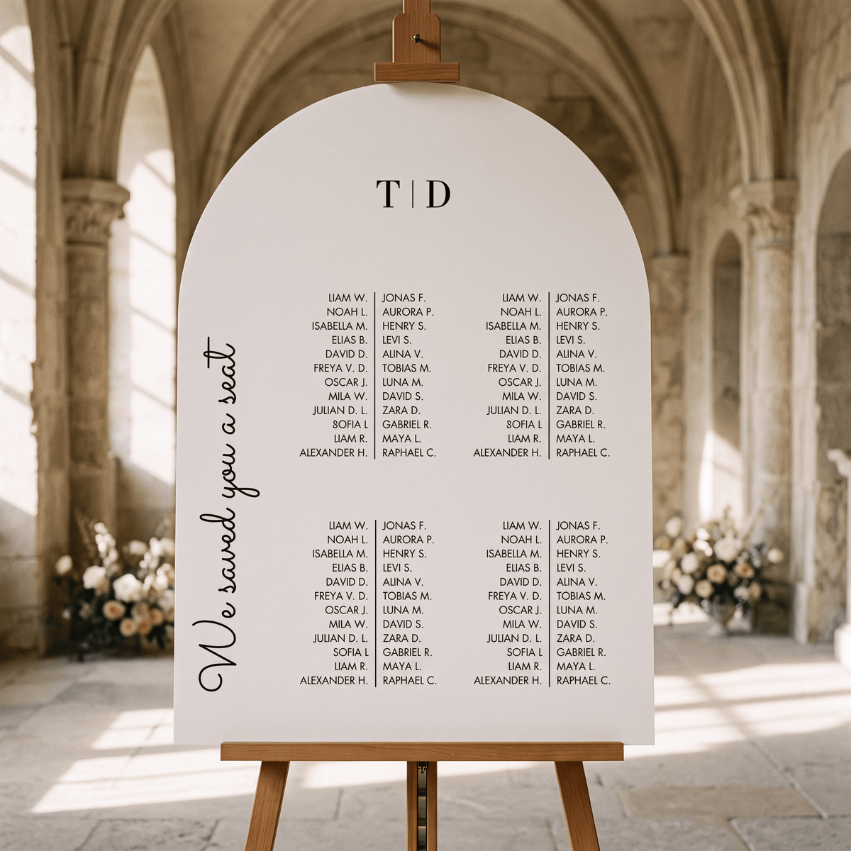 A large white arch-shaped wedding seating sign displayed on a wooden easel in a bright, sunlit hall. The sign features the text 'We saved you a seat' on the left, 'TOD' at the top center, and multiple columns of printed names.