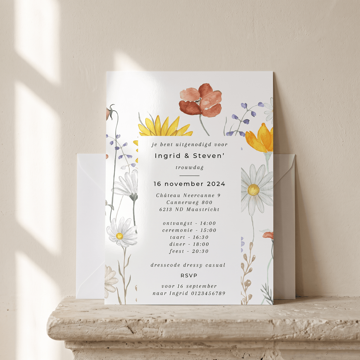 A vertical wedding invitation card with floral illustrations in orange, yellow, and white, displayed on a stone ledge with a matching envelope behind it against a warm beige wall.