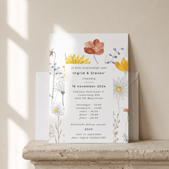 A vertical wedding invitation card with floral illustrations in orange, yellow, and white, displayed on a stone ledge with a matching envelope behind it against a warm beige wall.