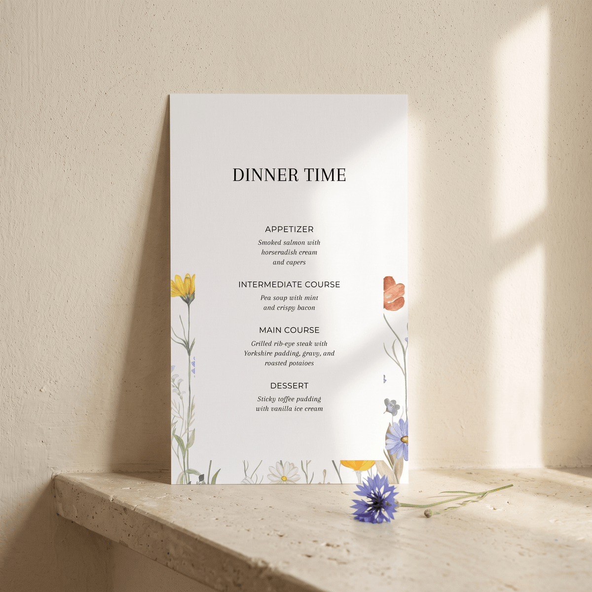 A vertical dinner time menu card with elegant black typography and delicate floral illustrations, leaning against a textured beige wall on a stone ledge with soft sunlight casting shadows.
