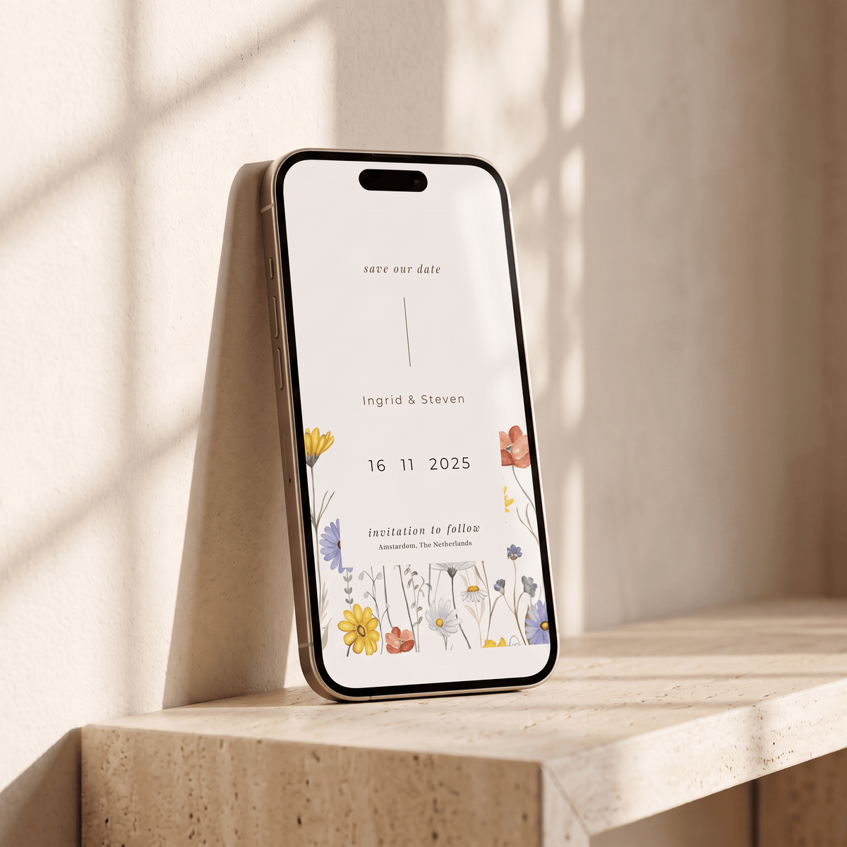 A smartphone propped upright on a stone surface against a warm beige wall, showing a floral 'save the date' wedding invitation design on the screen.