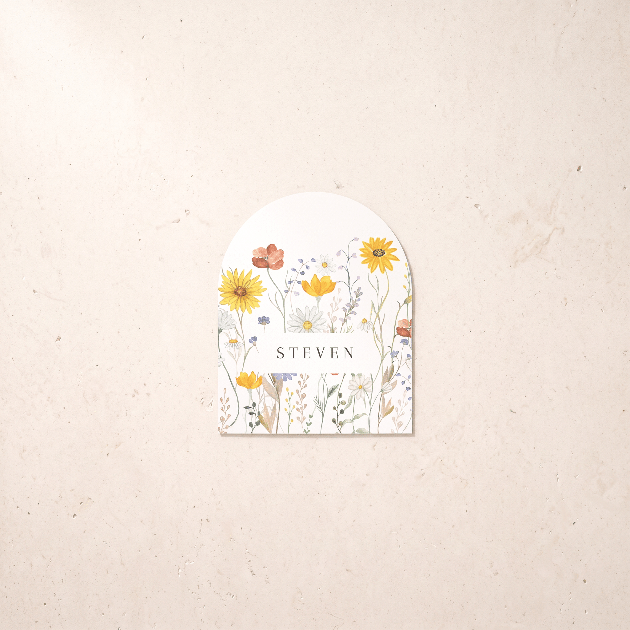 A personalized arched paper tag or label with a watercolor wildflower design and the name 'STEVEN' in the center, displayed on a textured beige background.