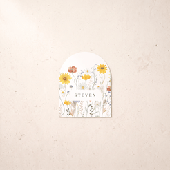 A personalized arched paper tag or label with a watercolor wildflower design and the name 'STEVEN' in the center, displayed on a textured beige background.