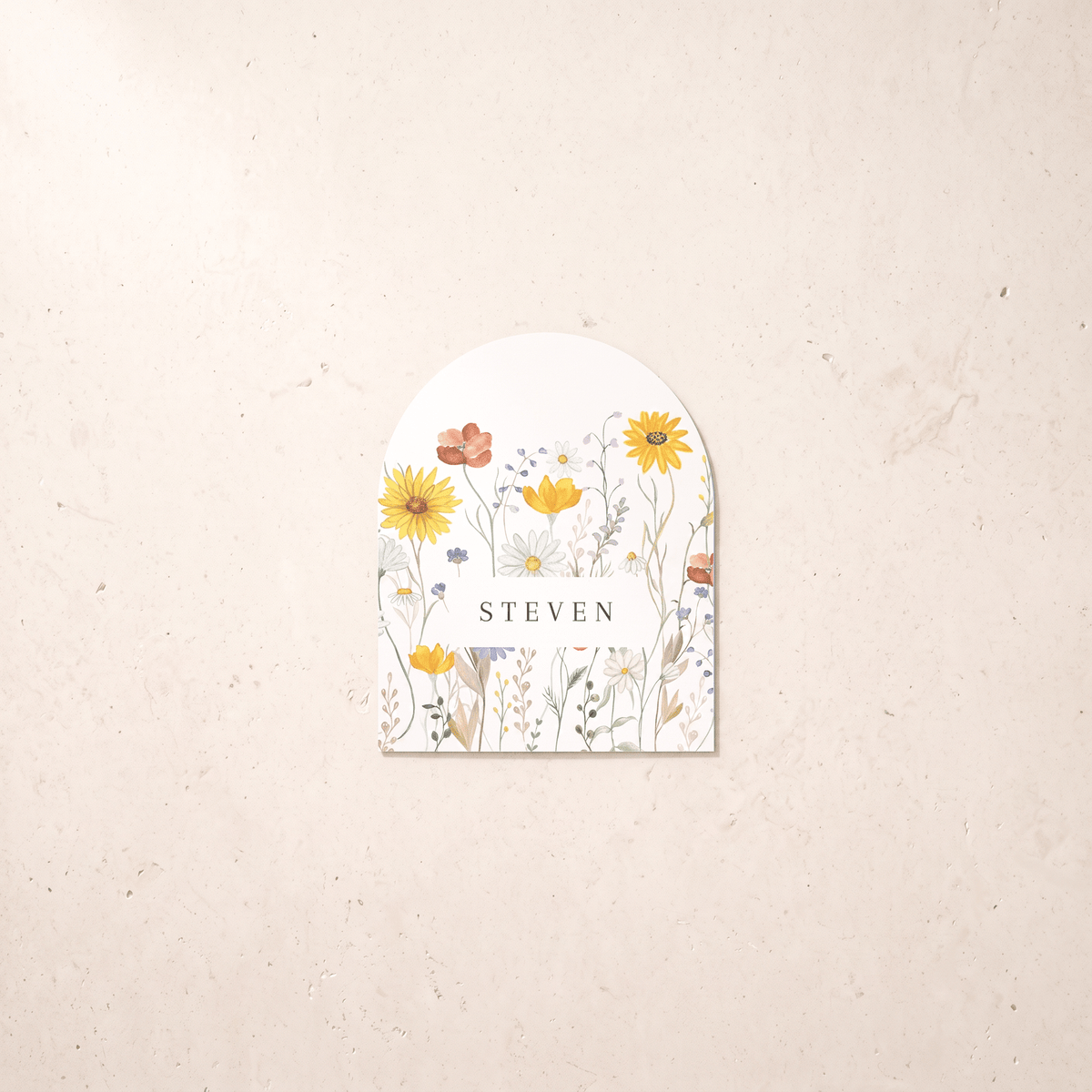 A personalized arched paper tag or label with a watercolor wildflower design and the name 'STEVEN' in the center, displayed on a textured beige background.