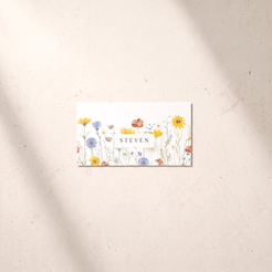 A rectangular white card featuring a colorful watercolor wildflower pattern and the name 'STEVEN' in the center, placed on a light beige textured surface with soft diagonal shadows.