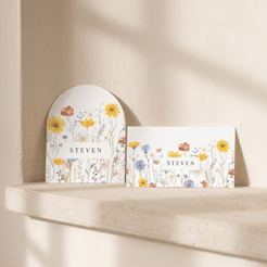 Two white floral greeting cards with the name 'STEVEN' printed in the center, displayed on a light stone ledge against a textured beige wall with soft sunlight shadows.