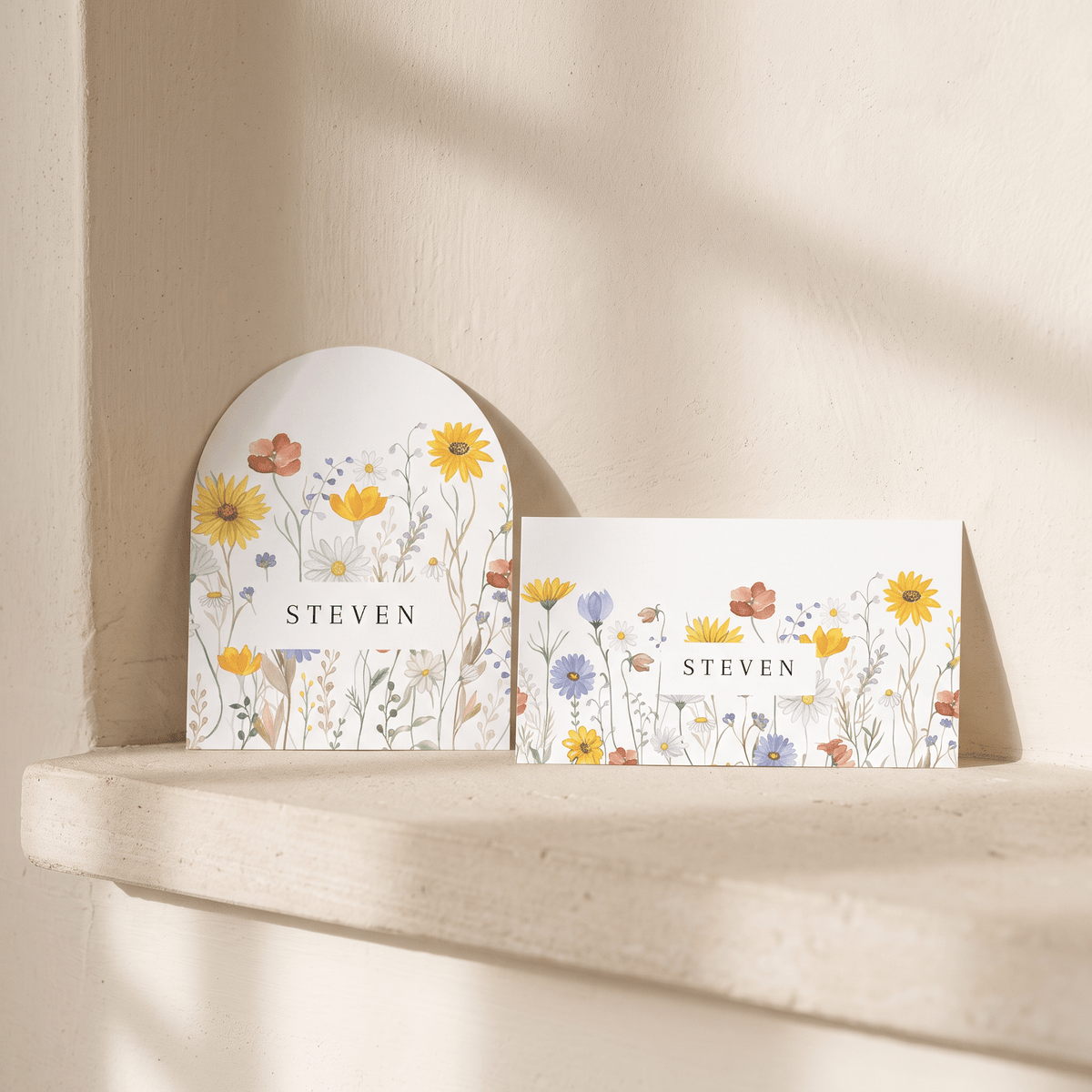 Two white floral greeting cards with the name 'STEVEN' printed in the center, displayed on a light stone ledge against a textured beige wall with soft sunlight shadows.