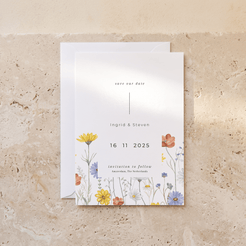 A minimalist wedding save-the-date card with a white background and delicate watercolor floral illustrations in yellow, orange, blue, and purple, displayed on a textured beige stone surface with a white envelope behind it.