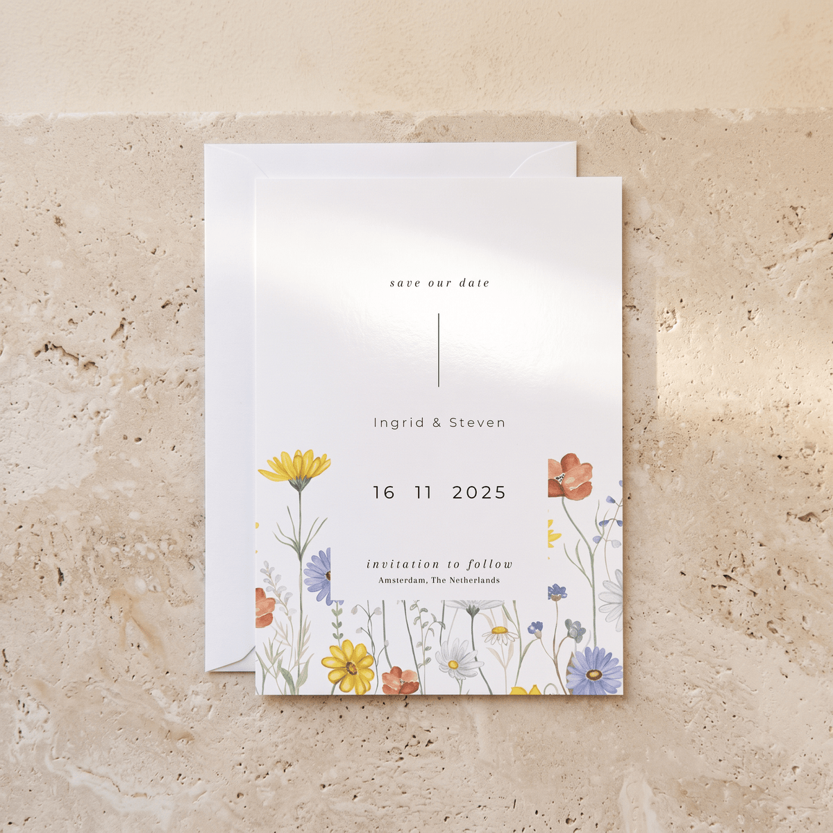 A minimalist wedding save-the-date card with a white background and delicate watercolor floral illustrations in yellow, orange, blue, and purple, displayed on a textured beige stone surface with a white envelope behind it.