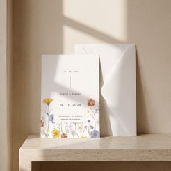 A white wedding save-the-date card with delicate multicolor wildflower illustrations at the bottom, standing on a stone ledge next to a white envelope against a warm beige wall with soft window shadows.