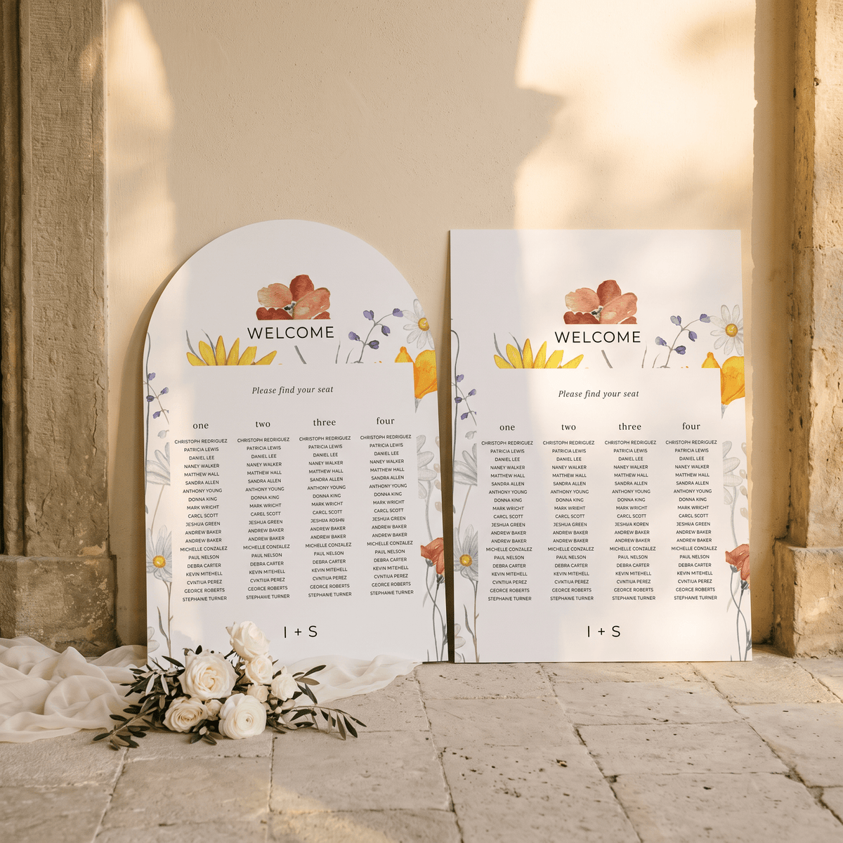Two floral wedding seating charts, one arched and one rectangular, displayed on a stone floor against a beige wall with sunlight and shadows.