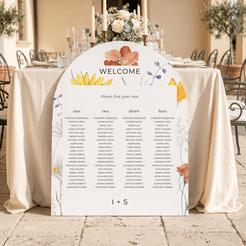 A floral wedding seating chart sign with a 'WELCOME' header and guest names organized in columns, displayed on a decorated outdoor event table with white tablecloths and glassware.