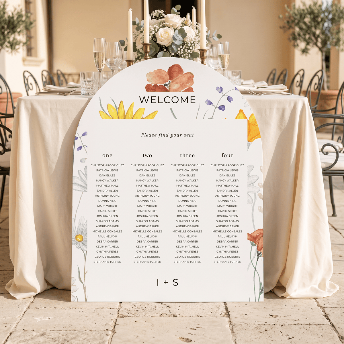 A floral wedding seating chart sign with a 'WELCOME' header and guest names organized in columns, displayed on a decorated outdoor event table with white tablecloths and glassware.