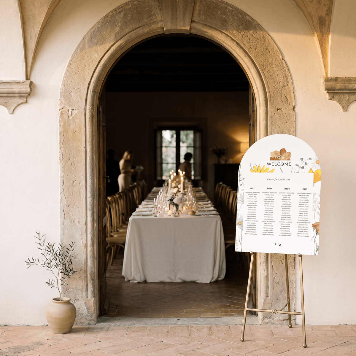 A wedding welcome sign on a gold easel in front of a stone archway, looking into a beautifully set dining room with a long table, chairs, and candles. A small potted plant sits on the floor to the left.