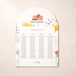 A printed welcome seating chart with floral watercolor illustrations around the border, shown on a light beige textured background. The card includes sections for one through four tables and names under a large 'WELCOME' heading.