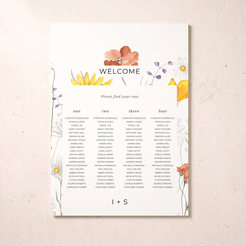 A printed wedding welcome seating card with floral watercolor illustrations in yellow, orange, purple, and blue, displayed against a light beige textured background. The card includes columns for guests numbered one through four and the text 'WELCOME' and 'Please find your seat'.