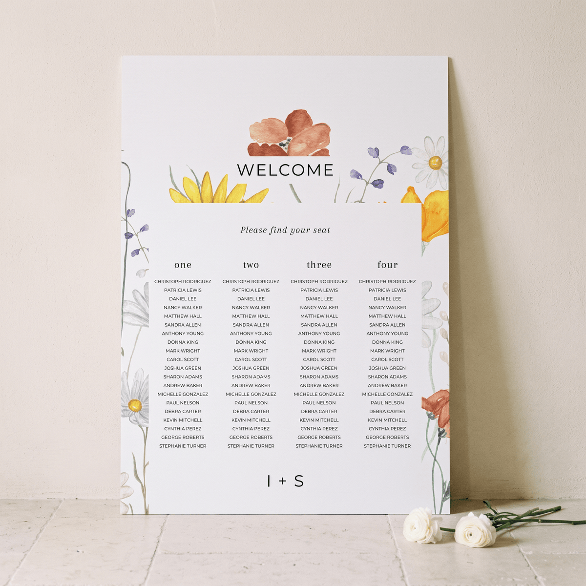 A printed welcome seating chart with floral watercolor illustrations in yellow, orange, purple, and green, displayed against a light beige wall with a few white roses on the floor.