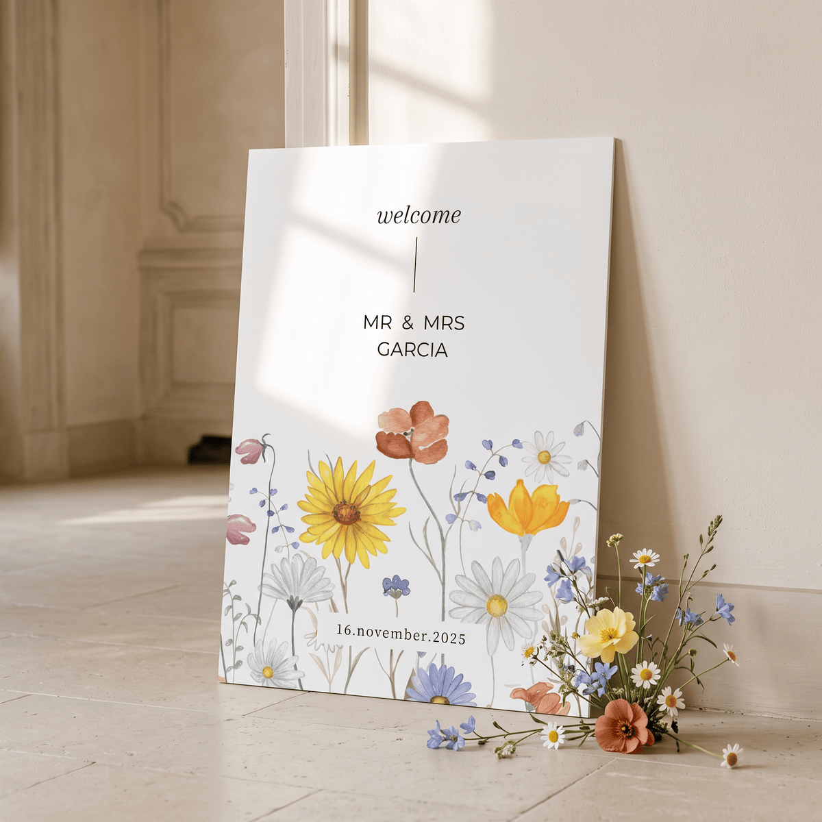 A vertical white wedding welcome sign leaning against a cream wall on a stone floor, printed with the words 'welcome', 'MR & MRS GARCIA', and '16 november 2025' above a watercolor floral arrangement of yellow, orange, blue, and white flowers. A matching loose flower bouquet rests beside the sign.