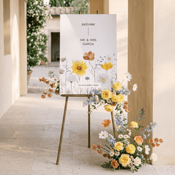 A vertical wedding welcome sign on a gold easel in a sunlit stone corridor, decorated with a floral arrangement of yellow, orange, and white flowers at the base. The sign reads 'welcome' and 'Mr.