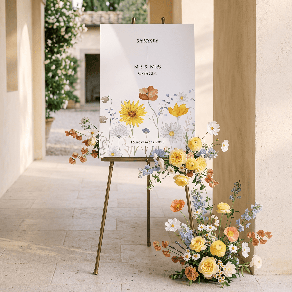 A vertical wedding welcome sign on a gold easel in a sunlit stone corridor, decorated with a floral arrangement of yellow, orange, and white flowers at the base. The sign reads 'welcome' and 'Mr.