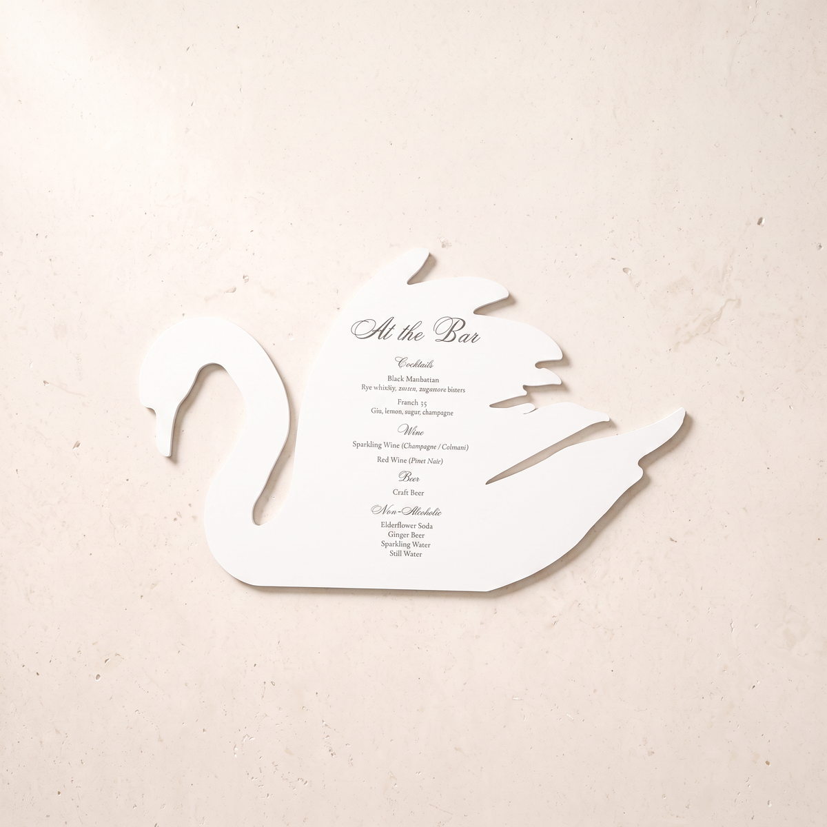 A white swan-shaped paper menu card with elegant black script and serif text reading 'At the Bar' and a list of course titles, placed on a light beige textured surface.