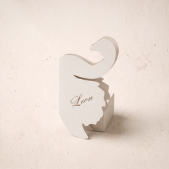 A white paper cutout place card shaped like a stylized letter B with the name 'Leon' printed in black script, standing on a textured beige surface.